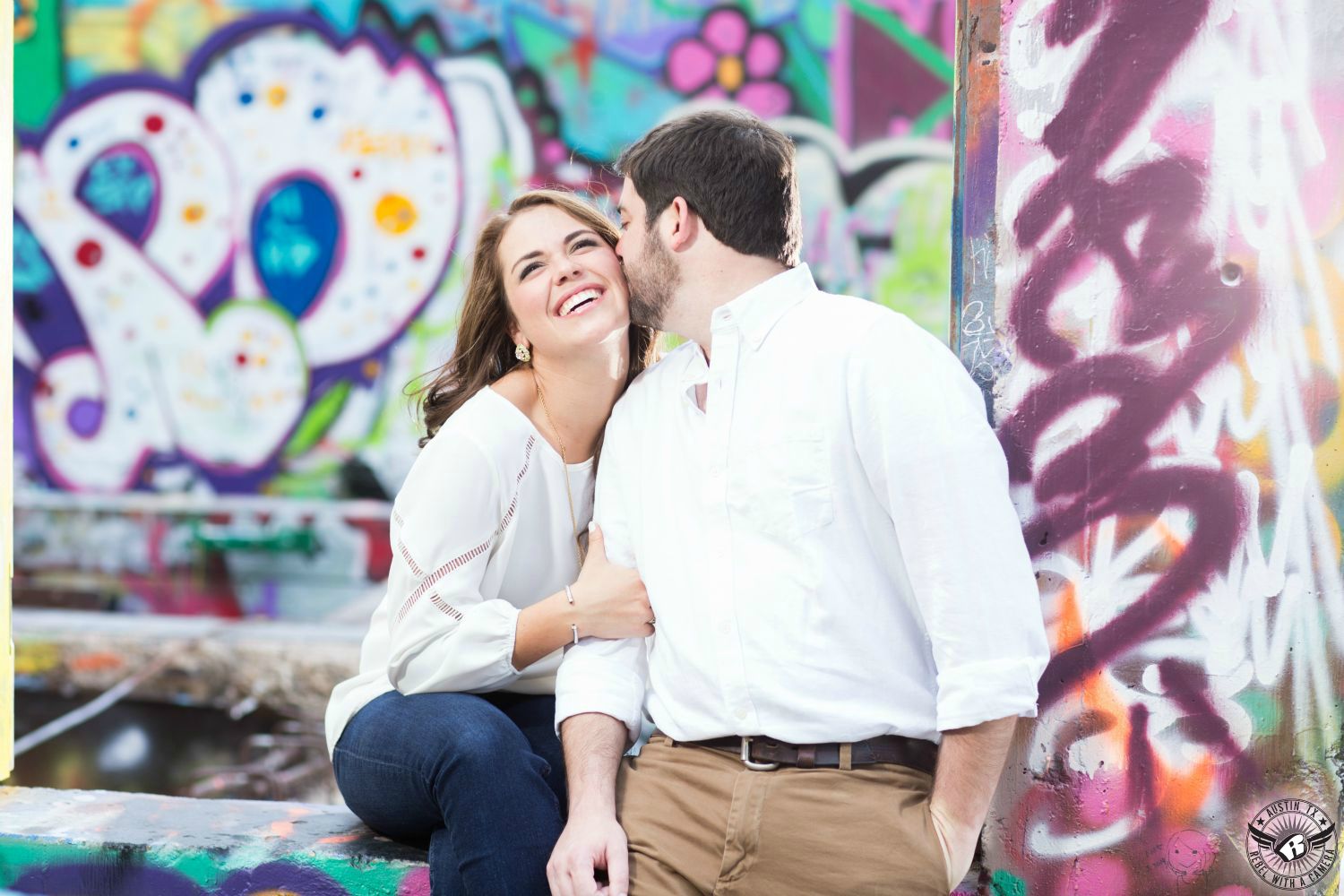 engagement of brunette girl  in white blouse  and blue jeans laughs as guy in white button up shirt in tan pants kisses her cheek at the castle hill graffiti park with lots of colorful painted art all over the wall in this fun engagement portrait